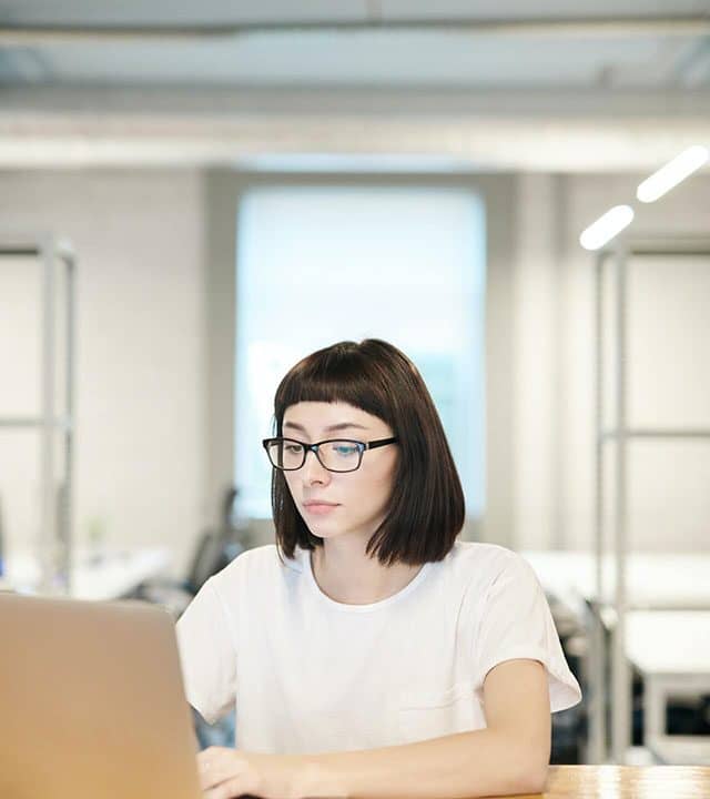 A confident and stylish woman wearing glasses is engrossed in her work on a laptop. Her determination and focus shine through as she works diligently, adding to the vibrant atmosphere of productivity in the coworking space.